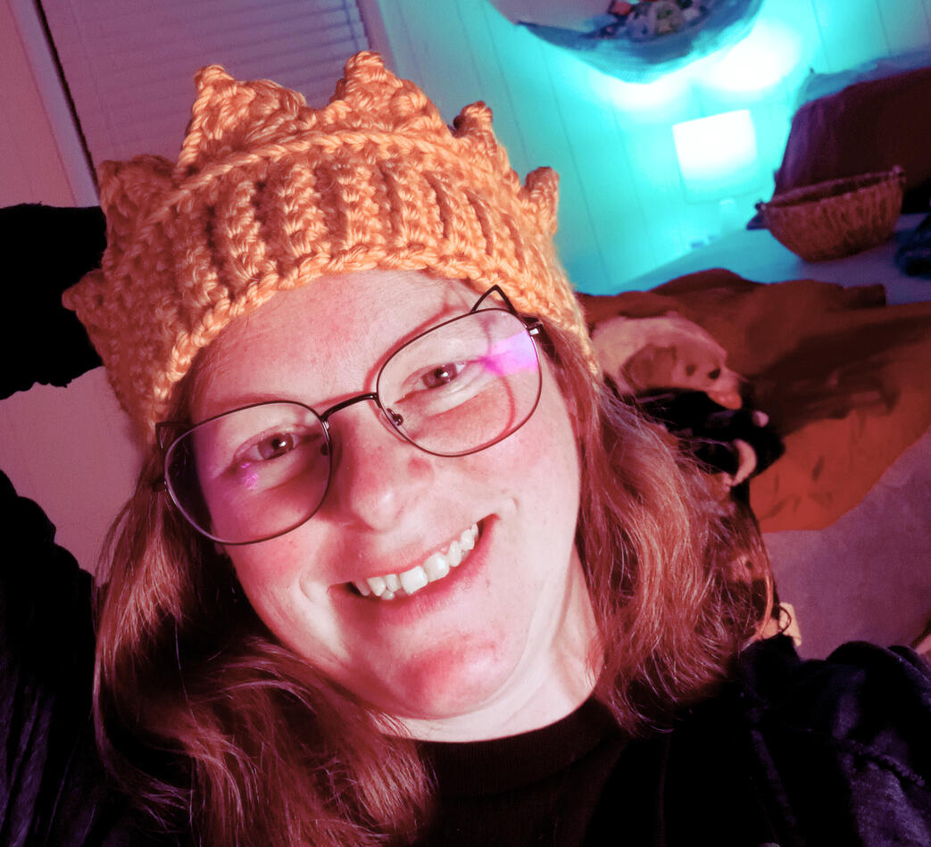 Laura | Lala Weiller, the site owner Laura Weiller, the owner and author of this site faces the camera. She is wearing a pair of glasses, the rims of which have cat ears on them. There's some partially crocheted, multi-colored yarn with stitch markers down the edge of it in her left hand, whi